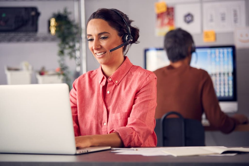 La imagen muestra a una mujer joven trabajando en una oficina moderna. Lleva auriculares con micrófono, lo que indica que está realizando tareas de atención al cliente o ventas por teléfono.